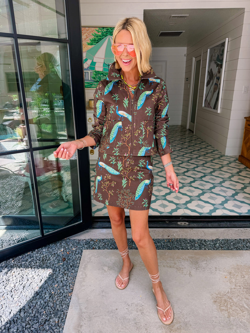 A woman wearing a brown denim zip-front jacket and matching mini skirt set with a vibrant blue and green peacock bird print.