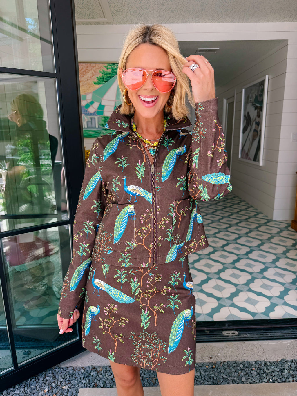 A woman wearing a brown denim zip-front jacket and matching mini skirt set with a vibrant blue and green peacock bird print.