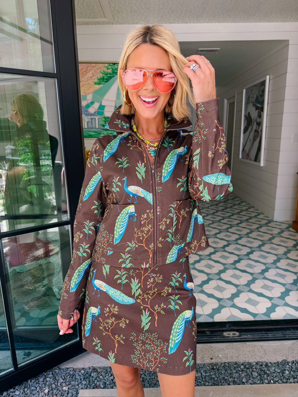 A woman wearing a brown denim zip-front jacket and matching mini skirt set with a vibrant blue and green peacock bird print.