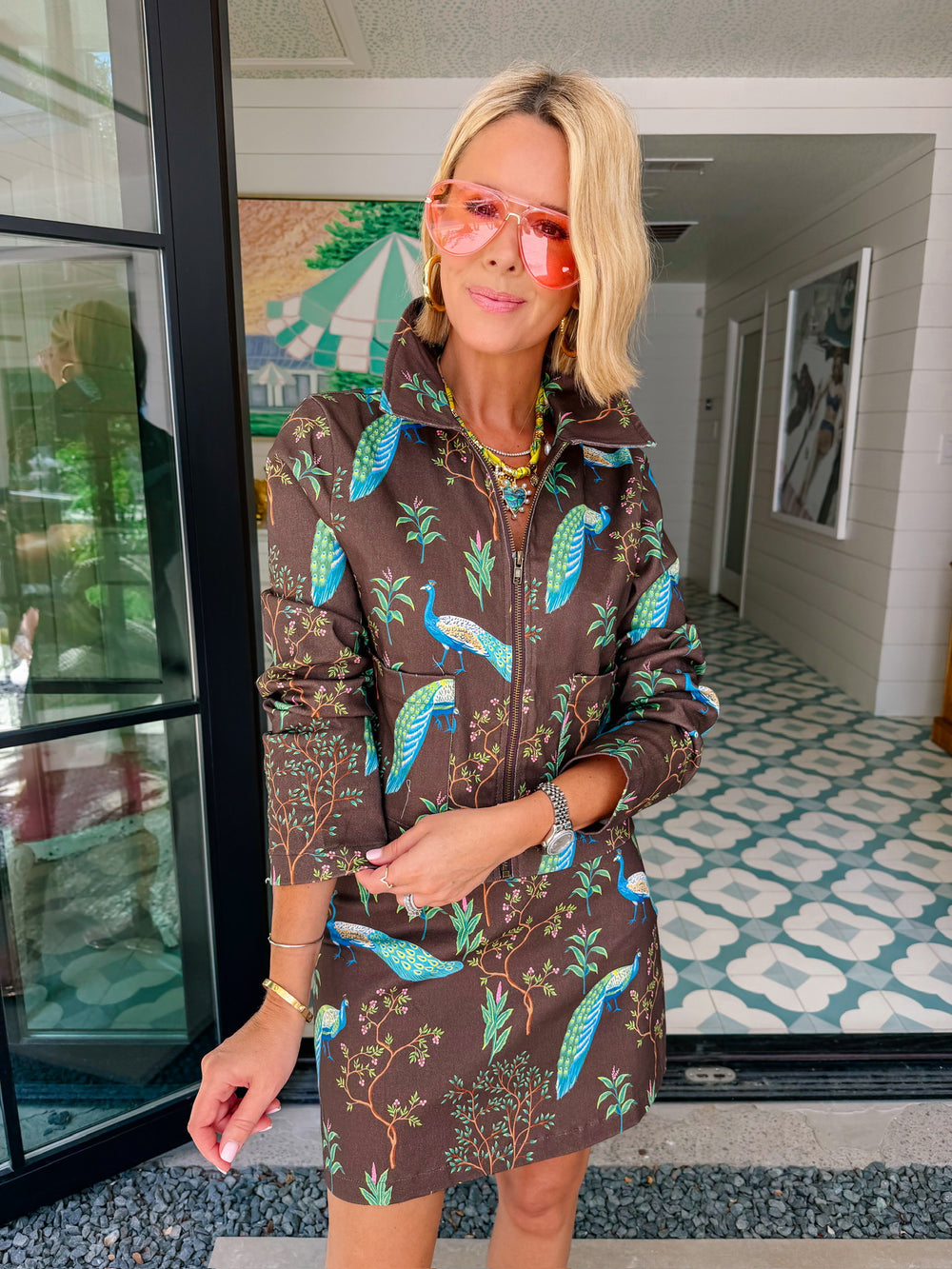 A woman wearing a brown denim zip-front jacket and matching mini skirt set with a vibrant blue and green peacock bird print.