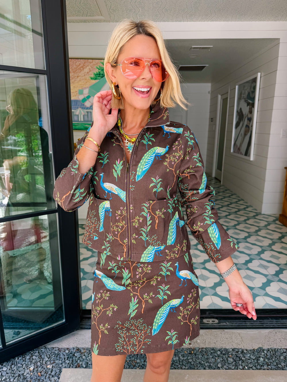 A woman wearing a brown denim zip-front jacket and matching mini skirt set with a vibrant blue and green peacock bird print.