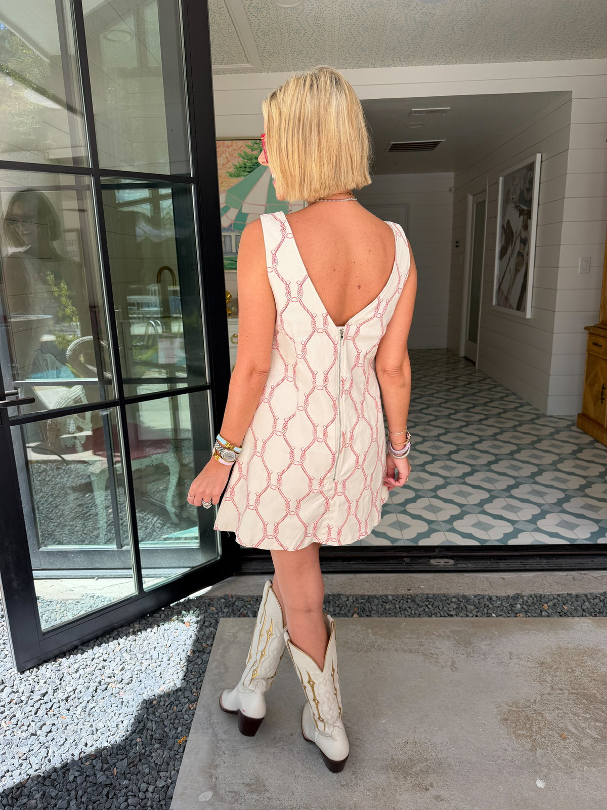 A woman wearing a sleeveless cream-colored A-line dress with a rodeo western motif pattern, styled with red sunglasses, a turquoise necklace, and bracelets. Wear to game day, rodeo or western events. SEC, game day, SMU, Texas Tech, TX Tech, OU, colts, NFL, rodeo, NFR, western