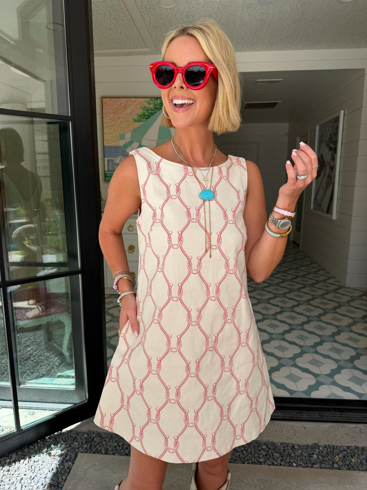 A woman wearing a sleeveless cream-colored A-line dress with a rodeo western motif pattern, styled with red sunglasses, a turquoise necklace, and bracelets. Wear to game day, rodeo or western events. SEC, game day, SMU, Texas Tech, TX Tech, OU, colts, NFL, rodeo, NFR, western