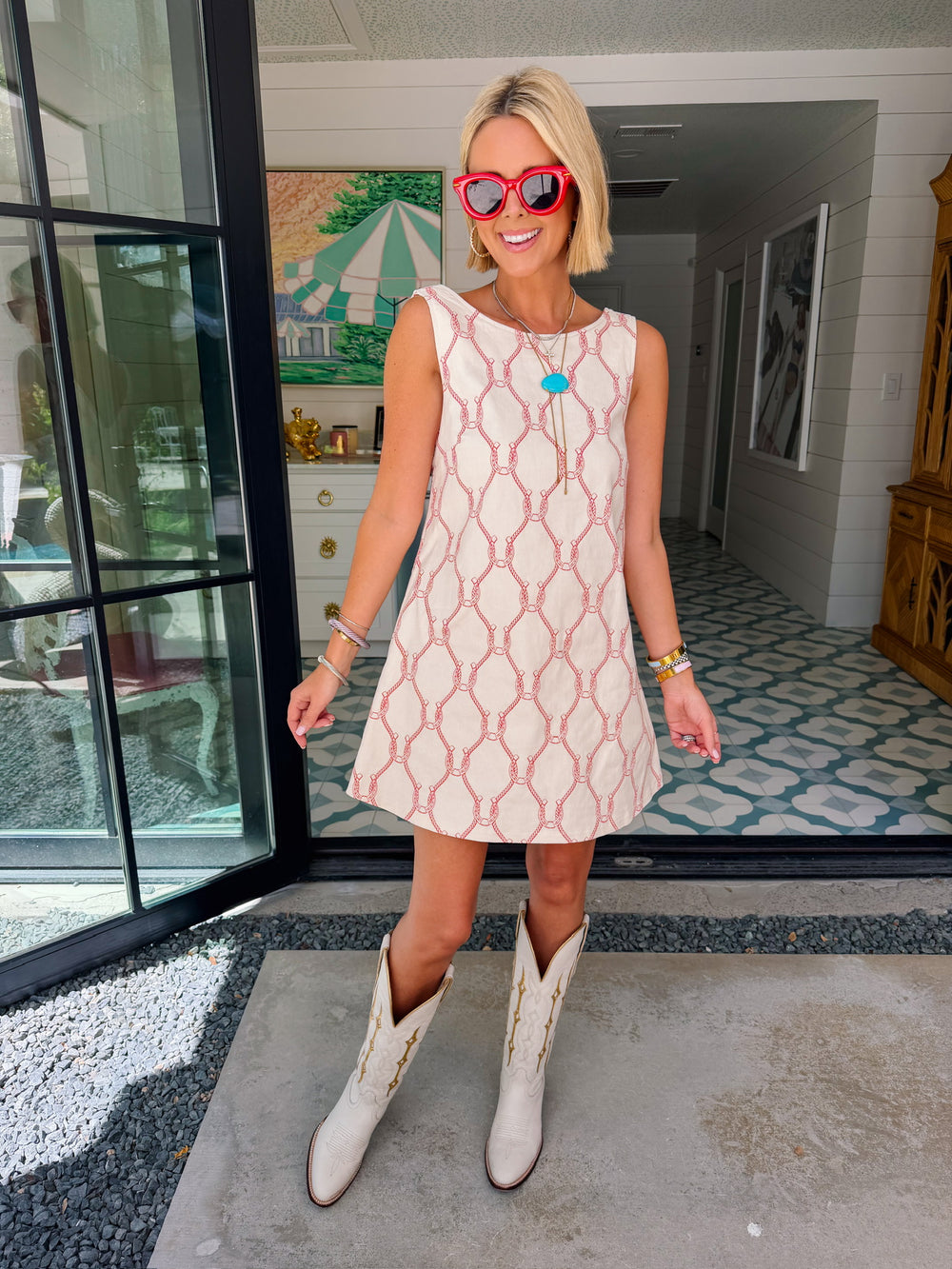 A woman wearing a sleeveless cream-colored A-line dress with a rodeo western motif pattern, styled with red sunglasses, a turquoise necklace, and bracelets. Wear to game day, rodeo or western events. SEC, game day, SMU, Texas Tech, TX Tech, OU, colts, NFL, rodeo, NFR, western