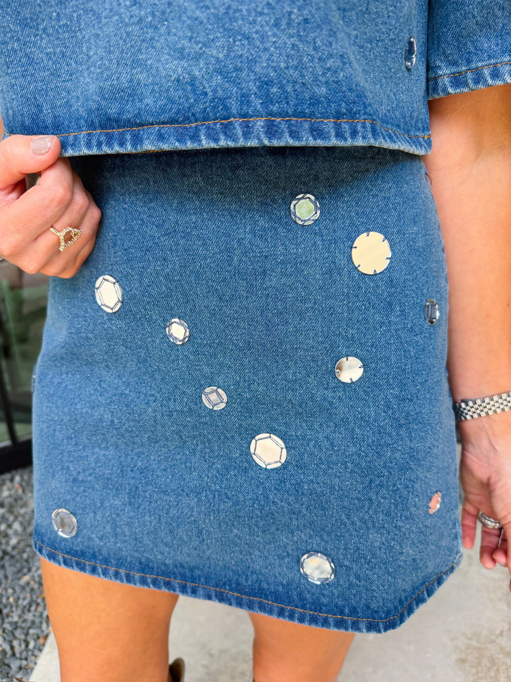 A woman modeling a blue denim short-sleeve top and matching mini skirt set, both decorated with mirror sequin embellishments. Wear this denim set to a concert, date night or gameday.