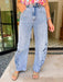 A women wearing light-wash wide-leg jeans with floral embroidery and eyelet cutout details on the outer thighs, paired with a colorful floral top and tan slide sandals. 