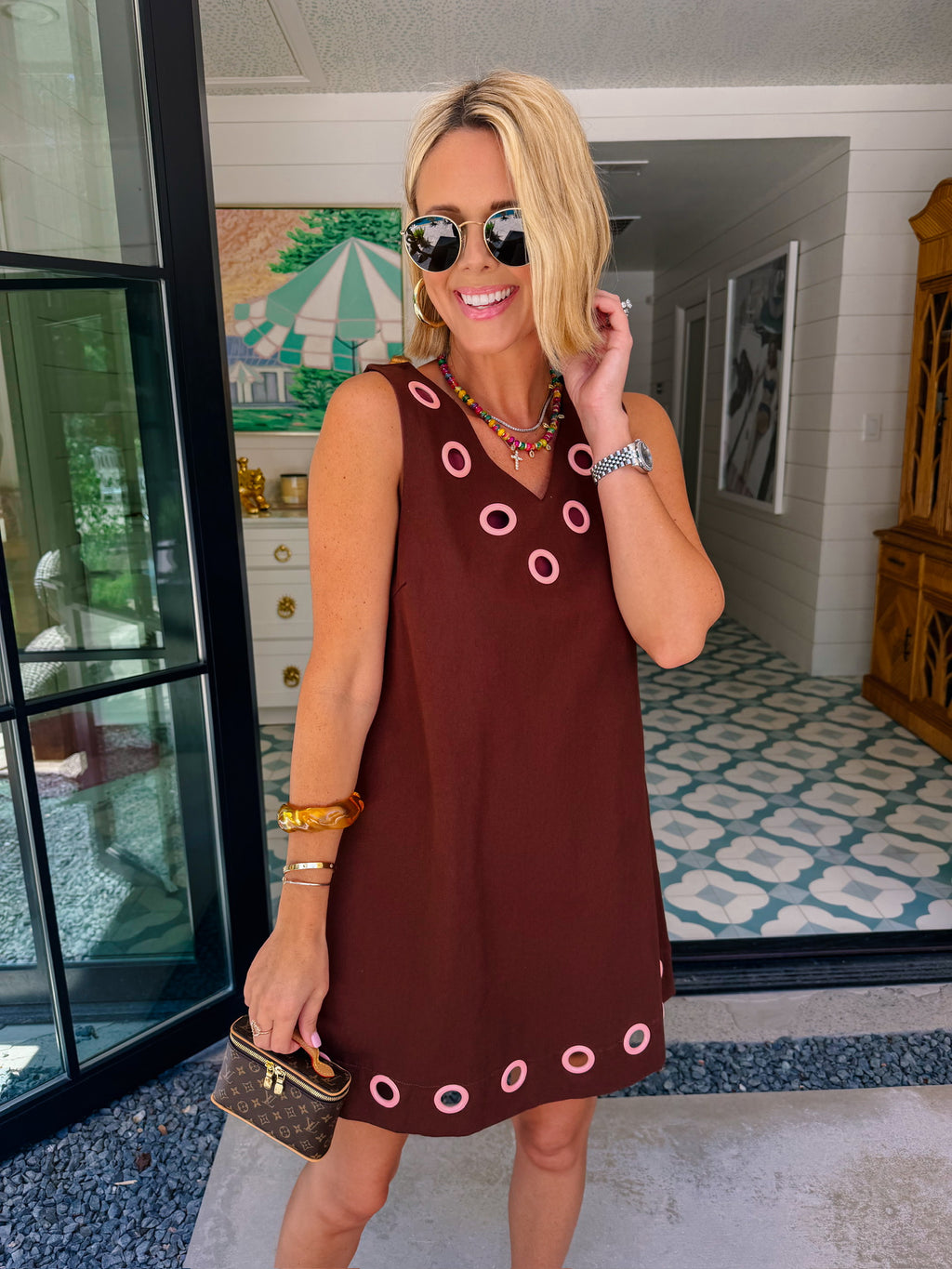 A woman modeling a sleeveless brown dress with circular cutout grommet details along neckline and hem.