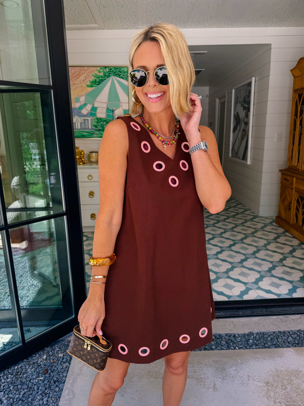 A woman modeling a sleeveless brown dress with circular cutout grommet details along neckline and hem.