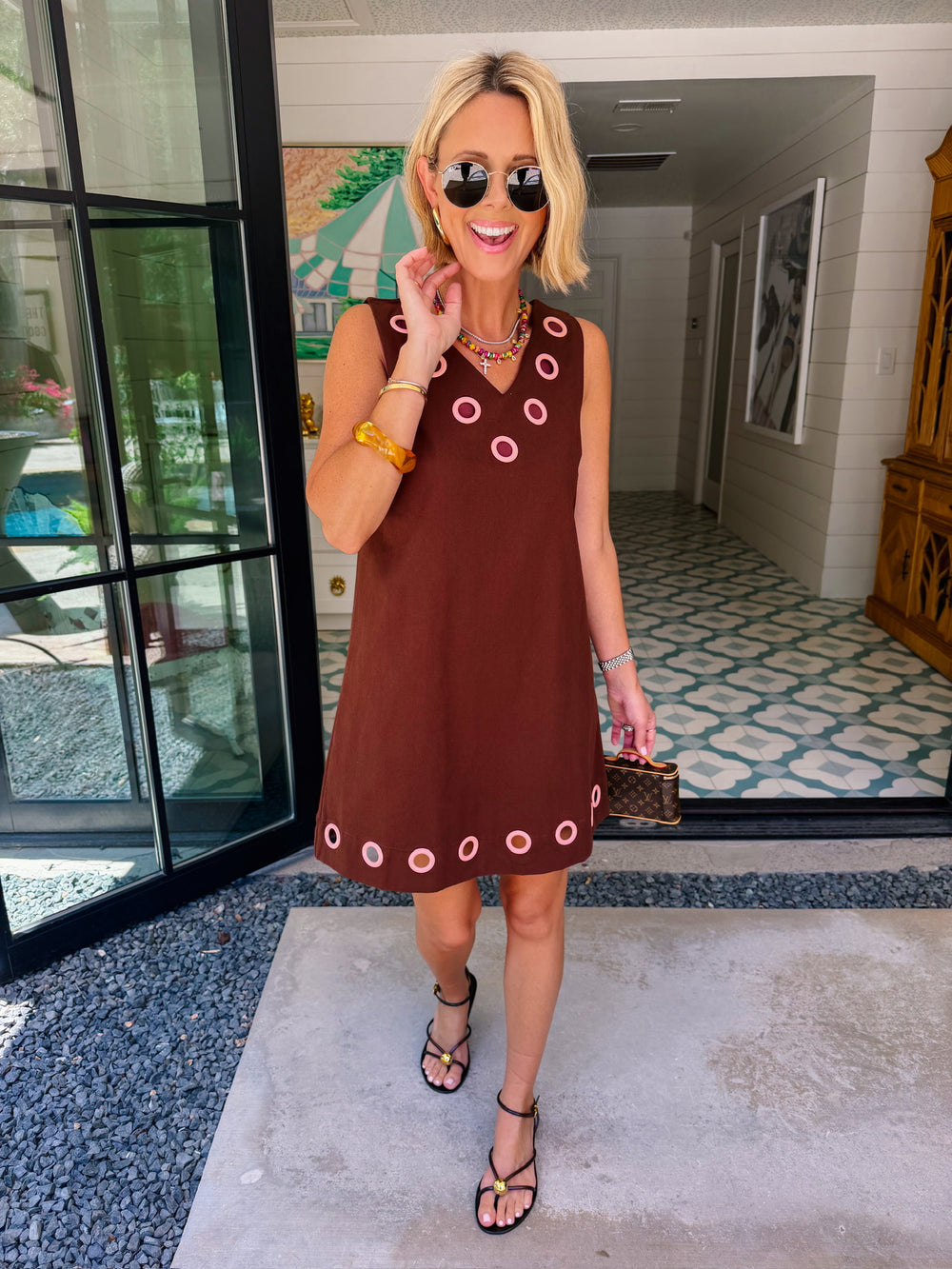 A woman modeling a sleeveless brown dress with circular cutout grommet details along neckline and hem.