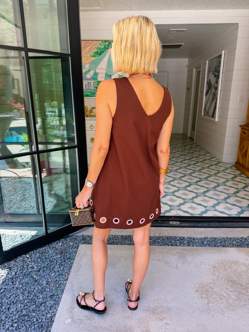 A women viewed from behind modeling a sleeveless brown dress with circular cutout grommet details along hem.