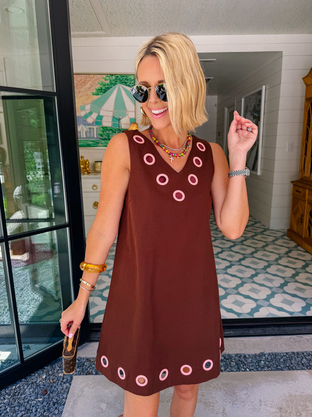 A woman modeling a sleeveless brown dress with circular cutout grommet details along neckline and hem.