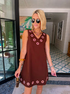 A woman modeling a sleeveless brown dress with circular cutout grommet details along neckline and hem.