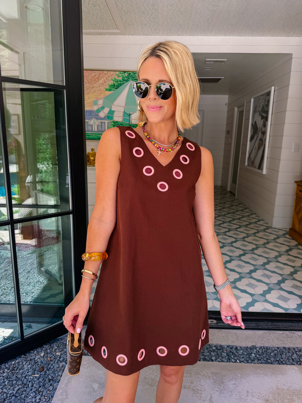 A woman modeling a sleeveless brown dress with circular cutout grommet details along neckline and hem.