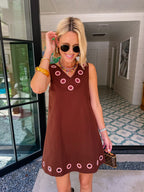 A woman modeling a sleeveless brown dress with circular cutout grommet details along neckline and hem.
