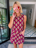 A woman wearing a sleeveless magenta mini dress with a yellow, blue, and white floral pattern, accessorized with pink-tinted sunglasses and a chunky gold necklace, standing in a home entryway.
