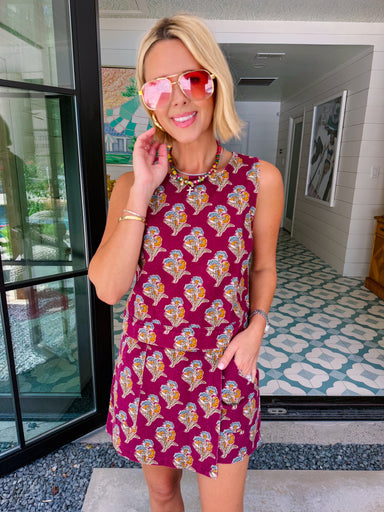 A woman wearing a sleeveless magenta mini dress with a yellow, blue, and white floral pattern, accessorized with pink-tinted sunglasses and a chunky gold necklace, standing in a home entryway.