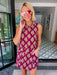 A woman wearing a sleeveless magenta mini dress with a yellow, blue, and white floral pattern, accessorized with pink-tinted sunglasses and a chunky gold necklace, standing in a home entryway.