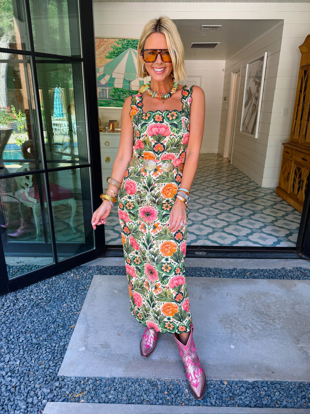 A woman wearing a sleeveless green maxi dress with a colorful floral print, accessorized with pink cowboy boots, sunglasses, and bracelets. The rich floral print combines green, peach and pink, making this a great transition piece for summer and fall.

