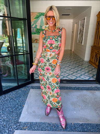 A woman wearing a sleeveless green maxi dress with a colorful floral print, accessorized with pink cowboy boots, sunglasses, and bracelets. The rich floral print combines green, peach and pink, making this a great transition piece for summer and fall.

