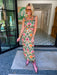 A woman wearing a sleeveless green maxi dress with a colorful floral print, accessorized with pink cowboy boots, sunglasses, and bracelets. The rich floral print combines green, peach and pink, making this a great transition piece for summer and fall.

