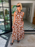 A woman stands in a home entryway wearing a sleeveless brown maxi dress with a white floral pattern. The dress features transition colors for summer and fall.

