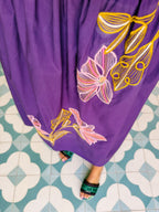 A woman wearing a purple spaghetti-strap midi dress with pink and green floral embroidery. Vacation look, TCU tailgate, gameday, Cabo, Mexico, beach, getaway