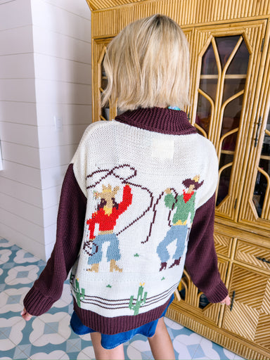 A woman wearing a western, rodeo themed cardigan sweater with brown sleeves. This cardi features cowboys and cowgirls lassoing, doing rope tricks. Perfect for rodeo, FW stockyards, stock show or any western event. Style with denim or over any dress.