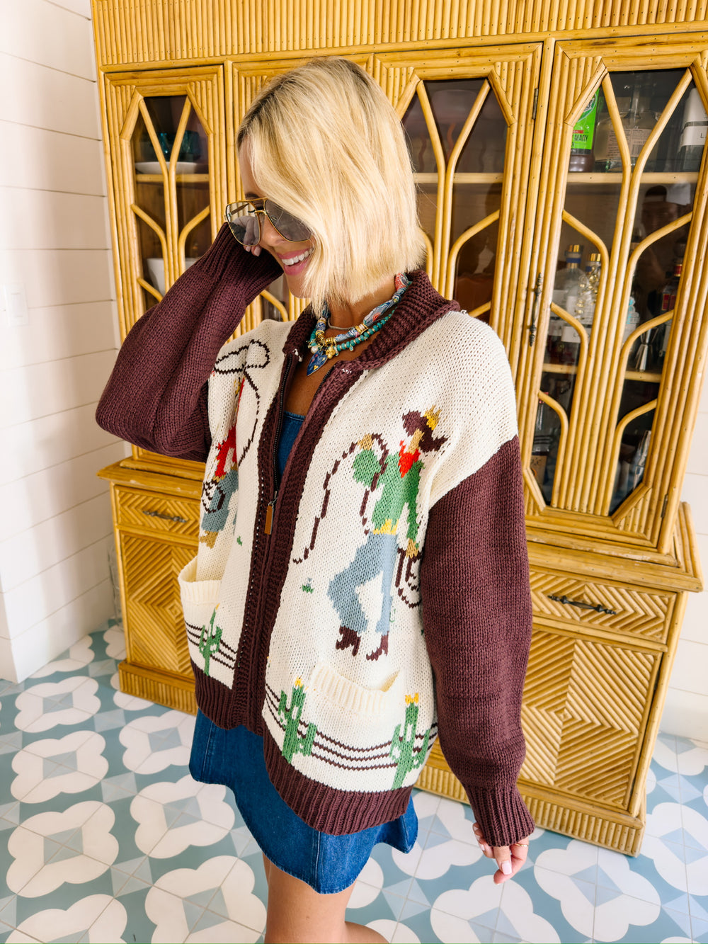 A woman wearing a western, rodeo themed cardigan sweater with brown sleeves. This cardi features cowboys and cowgirls lassoing, doing rope tricks. Perfect for rodeo, FW stockyards, stock show or any western event. Style with denim or over any dress.