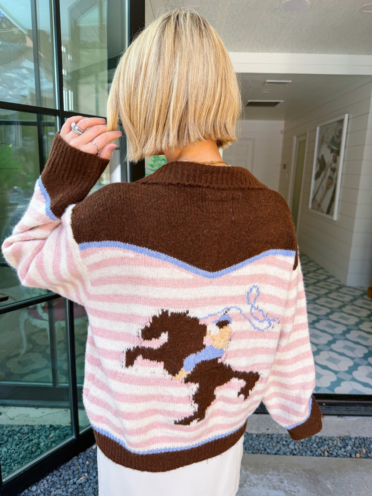 A woman wearing a western cardigan featuring a bronco and cowboy design on the back. This pink and white striped cardigan is perfect for rodeos, western events or country music concerts. Pair with denim or leather skirts.